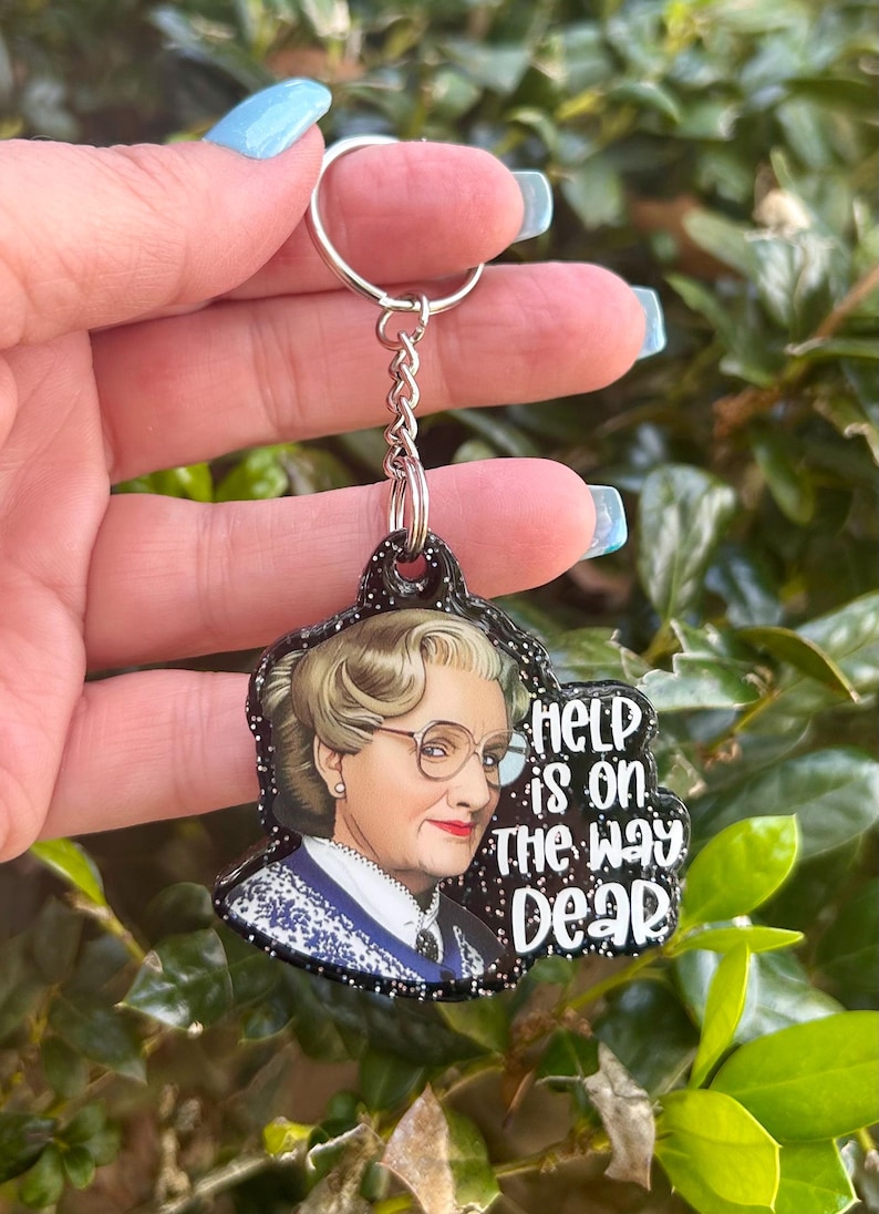 Puede incluir: Un llavero de purpurina negra con una imagen de una mujer con cabello rubio y gafas. El texto en el llavero dice "Help is on the way dear."