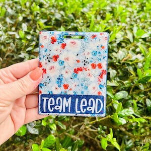 May include: A rectangular badge holder with a floral pattern of red, blue, and pink flowers on a light blue background. The bottom of the badge holder has the text "TEAM LEAD" in white letters on a blue glitter background.