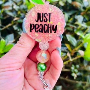 May include: A pink peach-shaped badge holder with the text "Just Peachy" in black lettering. The holder has a clear plastic strap and a green and iridescent bead charm.