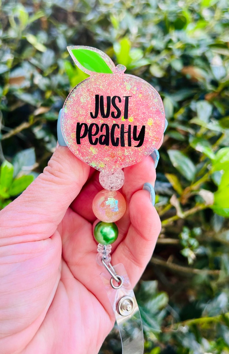 May include: A pink peach-shaped badge holder with the text "Just Peachy" in black lettering. The holder has a clear plastic strap and a green and iridescent bead charm.