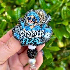 May include: A badge reel featuring a cartoon medical professional in blue scrubs, a cap, and sunglasses, making a rock-and-roll hand gesture. The design includes the text 'STERILE but FERAL' in white and blue, accented by black glitter and beads.