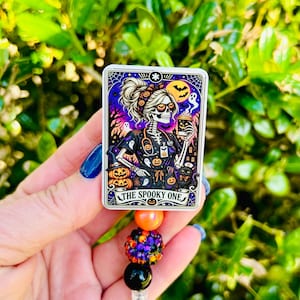 May include: A rectangular badge reel featuring a graphic of a skeleton wearing scrubs, holding a coffee cup, and surrounded by ghosts, pumpkins, and a black cat. The text reads THE SPOOKY ONE. Below the badge are three beads: orange, glittery purple-orange, and black.