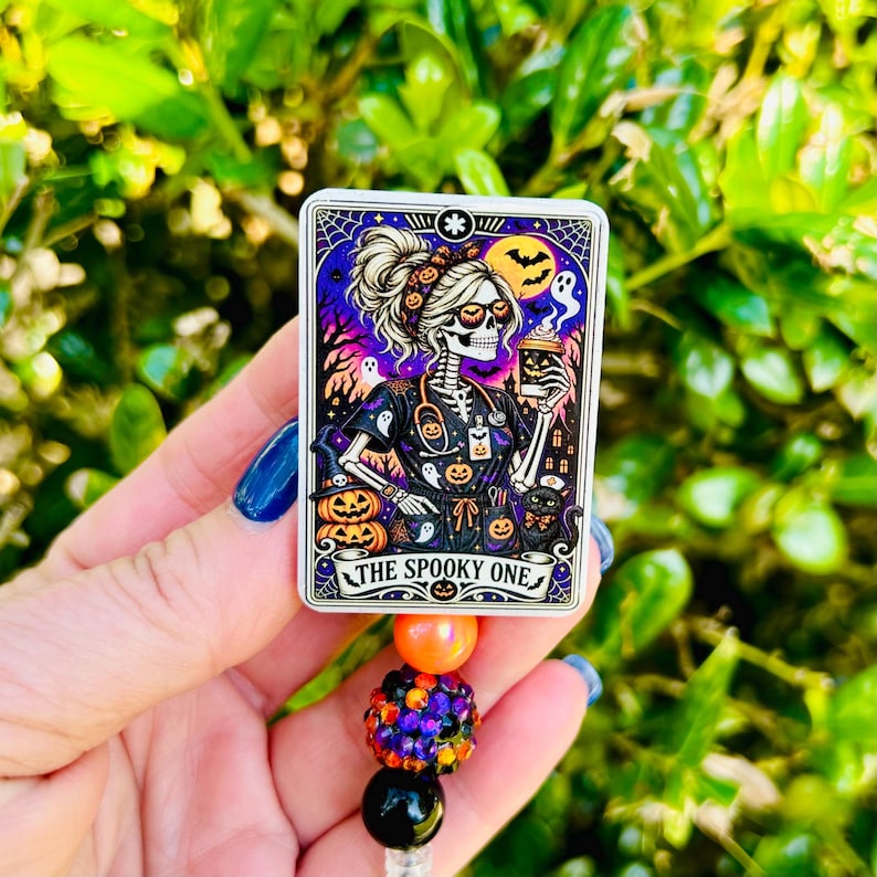 May include: A rectangular badge reel featuring a graphic of a skeleton wearing scrubs, holding a coffee cup, and surrounded by ghosts, pumpkins, and a black cat. The text reads THE SPOOKY ONE. Below the badge are three beads: orange, glittery purple-orange, and black.