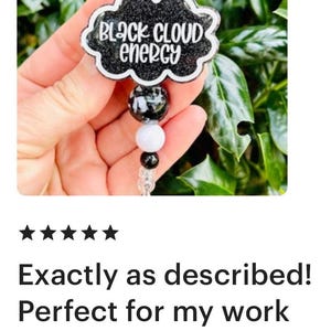 以下が含まれることがあります： 黒いグリッター仕上げの雲型バッジリール。白い文字で「BLACK CLOUD ENERGY」とスタイリッシュなフォントで書かれています。伸縮式のコードには、黒、白、透明の3つのビーズが装飾されており、先端にはプラスチック製のクリップが付いています。