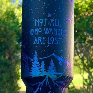 May include: A black stainless steel tumbler with a blue and green iridescent finish. The tumbler features a mountain scene with trees and a deer. The text "Not all who wander are lost" is printed on the tumbler.