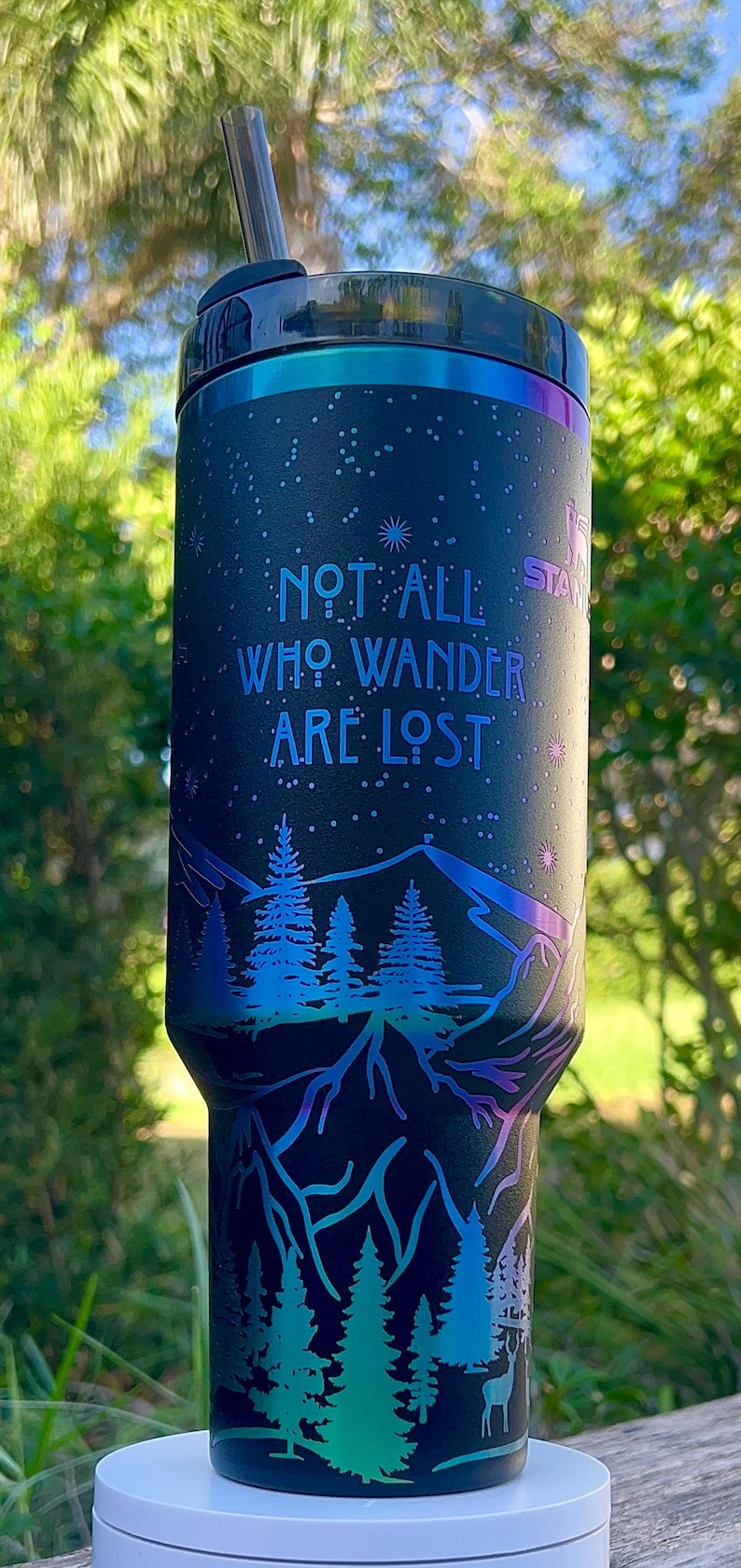 May include: A black stainless steel tumbler with a blue and green iridescent finish. The tumbler features a mountain scene with trees and a deer. The text "Not all who wander are lost" is printed on the tumbler.
