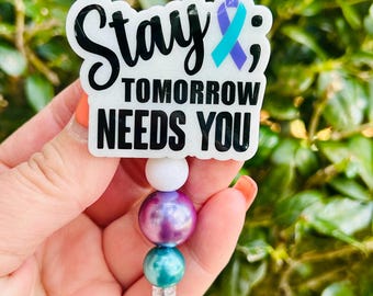 May include: A white badge with the words "Stay; Tomorrow Needs You" in black lettering. A teal and purple ribbon is on the right side. The badge is attached to a beaded lanyard with white, purple, and teal beads.