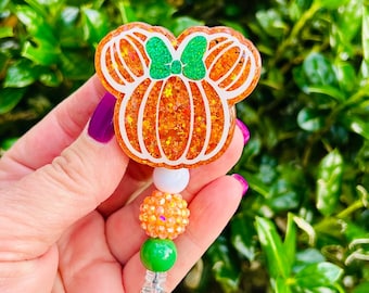 May include: A badge reel featuring a glittery orange pumpkin shaped like a mouse head with a green glitter bow. The pumpkin has white outlines and is attached to a vertical strand of beads including a white, an orange, and a green bead.