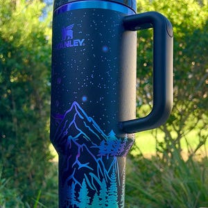 May include: A black Stanley travel mug with a starry night design and a mountain scene with trees and deer. The mug has a black handle and a silver lid.