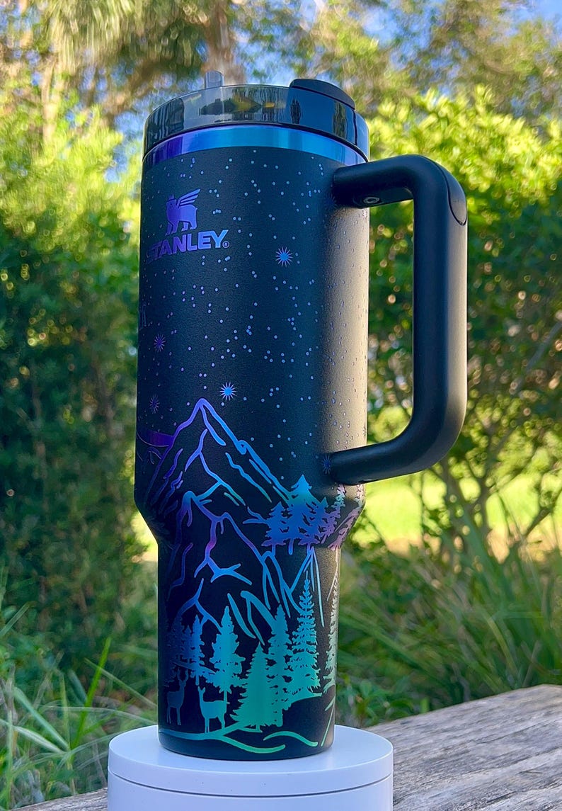 May include: A black Stanley travel mug with a starry night design and a mountain scene with trees and deer. The mug has a black handle and a silver lid.