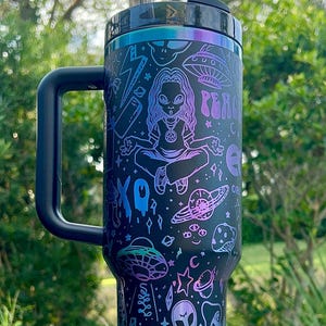 May include: Black tumbler with a handle, featuring a colorful design of an alien meditating, planets, UFOs, and the word "Groovy". The tumbler has a black lid and a black handle. The design includes purple, blue, and teal accents.