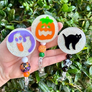 Puede incluir: Tres portatarjetas circulares con fondos brillantes. Uno muestra un fantasma morado, otro una calabaza naranja y el tercero la silueta de un gato negro. Cada carrete incluye un accesorio de cord&oacute;n con cuentas de colores variados.