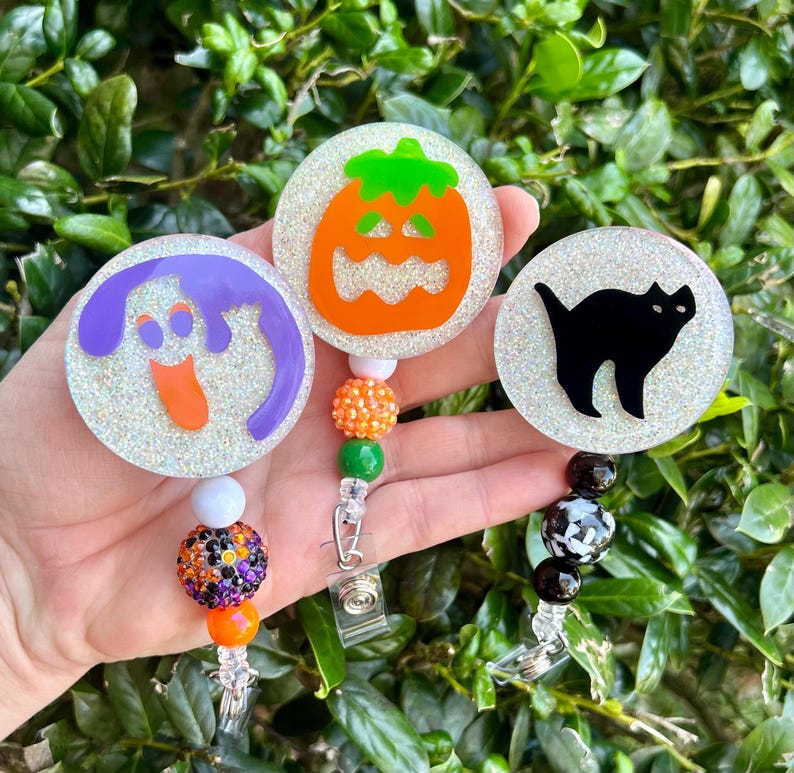 Puede incluir: Tres portatarjetas circulares con fondos brillantes. Uno muestra un fantasma morado, otro una calabaza naranja y el tercero la silueta de un gato negro. Cada carrete incluye un accesorio de cord&oacute;n con cuentas de colores variados.