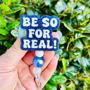 May include: Blue glitter badge reel with a white 'BE SO FOR REAL!' text overlay. The reel has a white bead and a blue iridescent bead.