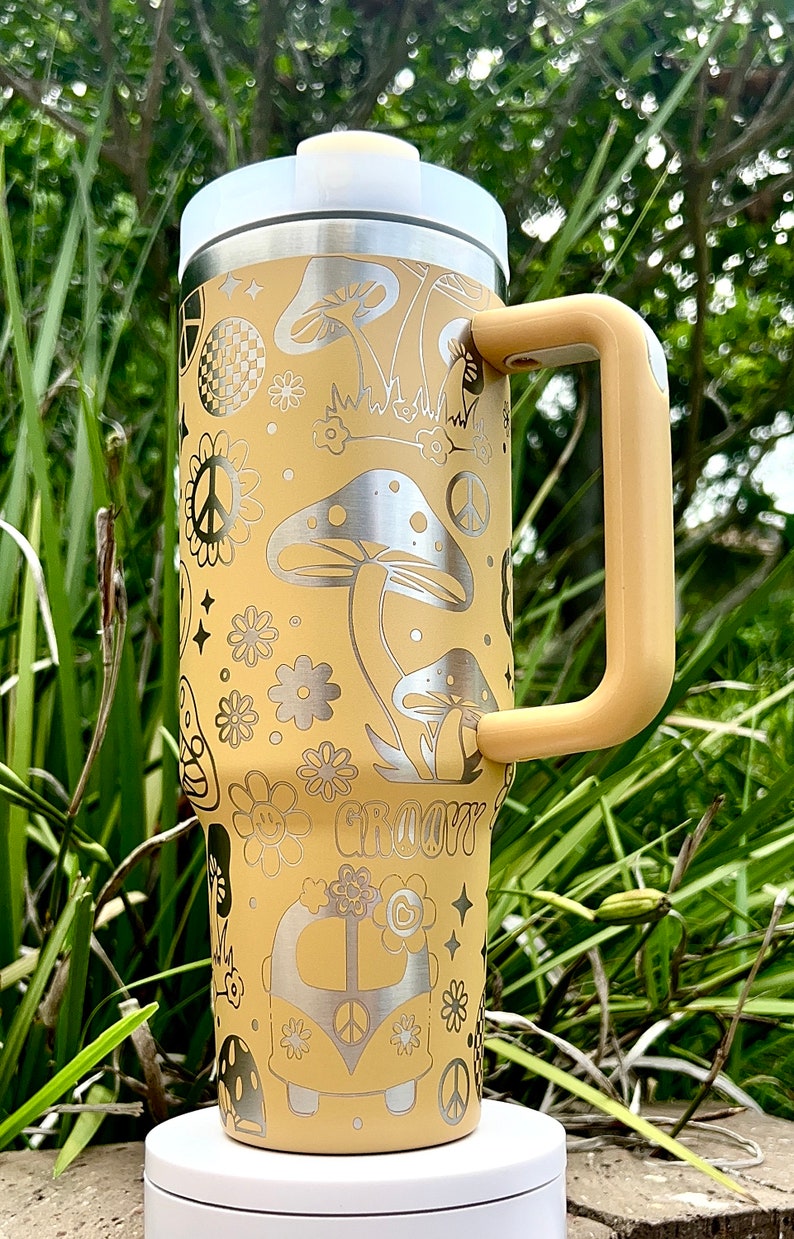 May include: A yellow stainless steel tumbler with a groovy 70s design featuring mushrooms, peace signs, flowers, and a vintage van. The tumbler has a lid and a handle.