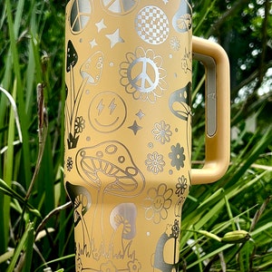 May include: A yellow stainless steel travel mug with a silver peace sign, mushrooms, and smiley faces. The mug has a handle and a lid.