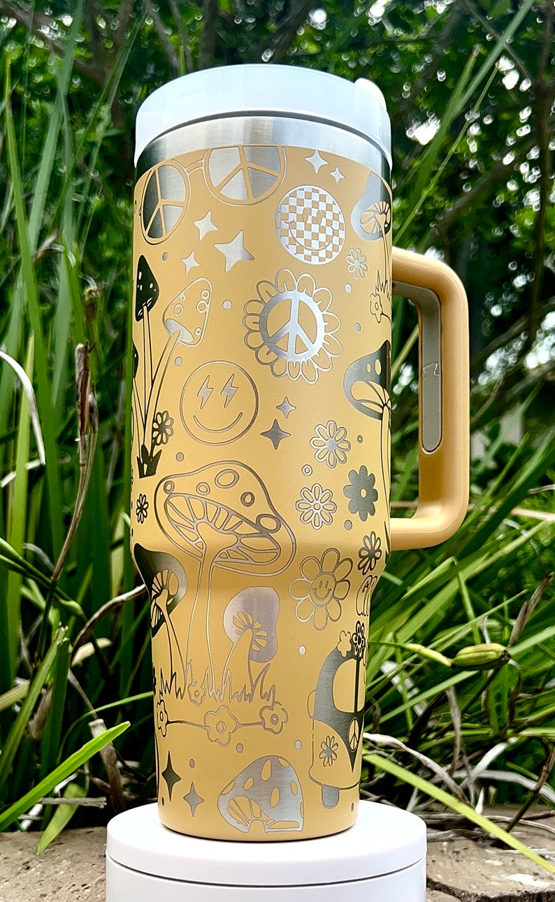 May include: A yellow stainless steel travel mug with a silver peace sign, mushrooms, and smiley faces. The mug has a handle and a lid.