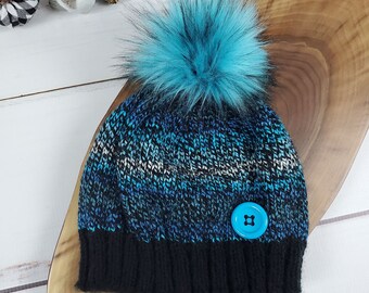 May include: A blue and black knit beanie with a fluffy blue pom pom and a blue button. The beanie is on a wooden surface.