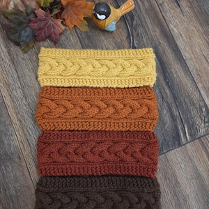 May include: A stack of four knitted headbands featuring a cable knit pattern. The headbands are colored in shades of mustard yellow, burnt orange, rust red, and dark brown, arranged vertically from lightest to darkest.