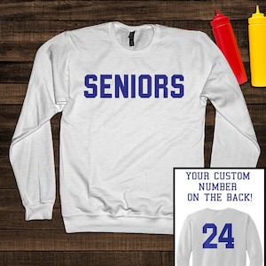 May include: A white long-sleeve crewneck sweatshirt featuring the word SENIORS in bold blue block letters across the chest. An inset shows the back of the sweatshirt with the number 24 in blue and the text YOUR CUSTOM NUMBER ON THE BACK!
