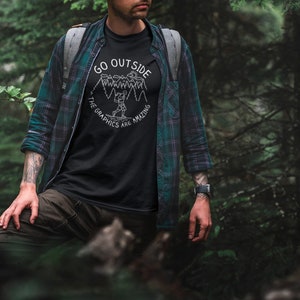 May include: A person wearing a black t-shirt with a white graphic that says "Go Outside The Graphics Are Amazing" and a green and black plaid shirt. The person is standing in a forest.