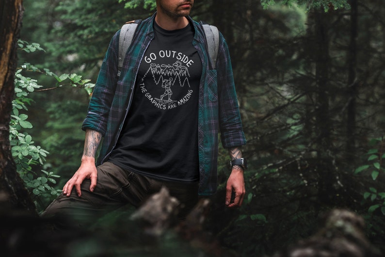 May include: A person wearing a black t-shirt with a white graphic that says "Go Outside The Graphics Are Amazing" and a green and black plaid shirt. The person is standing in a forest.