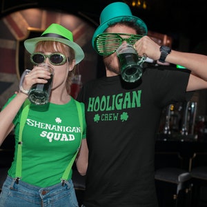 Puede incluir: Dos personas con camisas y sombreros verdes con dise&ntilde;os de tr&eacute;bol. La persona de la izquierda sostiene una jarra de cerveza verde y su camisa dice "Shenanigan Squad". La persona de la derecha sostiene una jarra de cerveza verde y su camisa dice "Hooligan Crew".