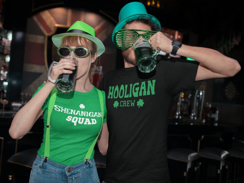 Puede incluir: Dos personas con camisas y sombreros verdes con dise&ntilde;os de tr&eacute;bol. La persona de la izquierda sostiene una jarra de cerveza verde y su camisa dice "Shenanigan Squad". La persona de la derecha sostiene una jarra de cerveza verde y su camisa dice "Hooligan Crew".