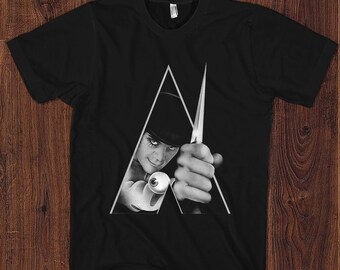 May include: Black and white graphic t-shirt featuring a character from the movie "A Clockwork Orange". The character is holding a knife and has a menacing expression. The image is in the shape of a triangle.