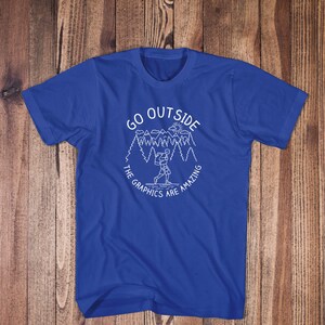 May include: A royal blue t-shirt with a white graphic design of a person hiking in a forest with the text "Go Outside The Graphics Are Amazing".