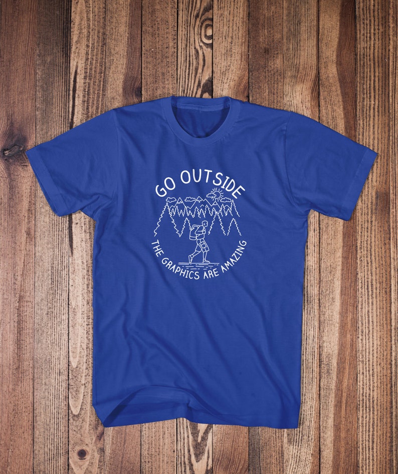May include: A royal blue t-shirt with a white graphic design of a person hiking in a forest with the text "Go Outside The Graphics Are Amazing".