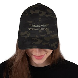 Op de afbeelding: Een camouflage baseball cap met de tekst "akashic. records" in een cursief lettertype. De pet heeft een gebogen klep en een gestructureerde pasvorm. De hoed heeft een donkergroen, bruin en zwart camouflagepatroon.