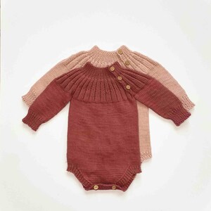 May include: Two knitted baby rompers. One is a deep red-brown color, and the other is a light pink. Both have long sleeves, a round neckline, and wooden buttons. The rompers are made of a soft, textured knit fabric.