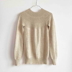 May include: A long-sleeved beige sweater featuring a ribbed knit yoke design around the neckline and shoulders. The garment has a crew neckline, fitted cuffs at the end of the sleeves, and a ribbed hemline, all rendered in a solid light tan color.