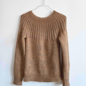 May include: A long-sleeved sweater in a solid tan color, featuring a round neckline with a ribbed knit pattern that extends across the upper chest and shoulders. The garment has a plain knit body and ribbed cuffs at the ends of the sleeves and hem.