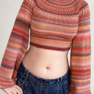 May include: A cropped, long-sleeved sweater with a ribbed neckline and cuffs. The sweater features horizontal stripes in shades of pink, orange, red, and purple. The sleeves are flared. The sweater is paired with dark blue denim jeans.