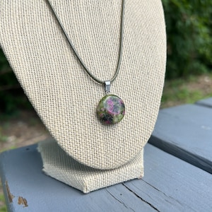 Peut inclure: Un collier avec un pendentif rond en pierre naturelle. La pierre présente des nuances de vert et de rose, sertie dans un métal argenté. Le collier a un cordon fin vert foncé. Le pendentif est présenté sur un tissu beige.