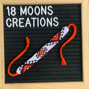 May include: A handmade friendship bracelet with a white, orange, and burgundy geometric pattern. The bracelet has orange braided ties on each end. The text "18 MOONS CREATIONS" is displayed in white block letters.