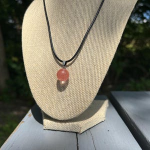 May include: A round, peach-colored gemstone pendant necklace. The pendant is suspended from a black cord and displayed on a neutral-toned jewelry stand. The background features a blurred outdoor scene with greenery.