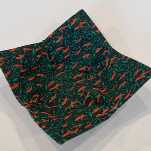 May include: A square fabric bowl with a black background, covered in a pattern of red chili peppers and green leaves. The bowl is folded to create a square shape, perfect for holding food.