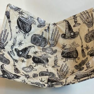 May include: A beige fabric bowl with a black Halloween-themed pattern. The pattern includes witch hats, cauldrons, pumpkins, skeleton hands, brooms, and spiders. The bowl is square-shaped with curved sides.