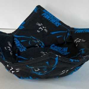 May include: A black fabric bowl with a blue and white Carolina Panthers logo and text. The bowl is folded into a square shape with pointed corners. The bowl is designed to hold food.