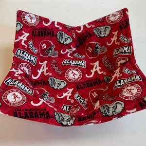 May include: A red and white fabric bowl cozy featuring a repeating pattern of the University of Alabama logos, including the letter "A", football helmets, and elephant mascots. The text "ALABAMA" and "BAMA" are also visible.