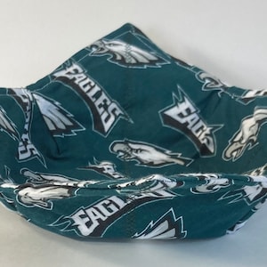 May include: A fabric bowl cozy featuring a repeating pattern of the Philadelphia Eagles logo and the word EAGLES in white, black, and silver on a dark teal background. The square-shaped holder has four corners and visible stitching.