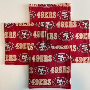 May include: A set of four square fabric coasters featuring a repeating pattern of the San Francisco 49ers logo and the text 49ERS in gold and white on a deep red background. Each coaster has stitched edges and a consistent, uniform design.