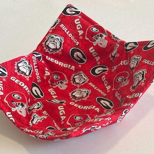 May include: A red fabric bowl cozy featuring a repeating pattern of Georgia Bulldogs logos, football helmets, and the text UGA and GEORGIA. The square-shaped quilted holder has four pointed corners and is constructed with visible stitching throughout.