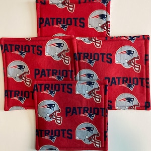 May include: A set of four square fabric coasters featuring a red background with a repeating pattern of grey football helmets and the word PATRIOTS printed in dark blue. Each coaster has stitched edges and a soft, quilted appearance.
