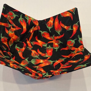 May include: A fabric bowl with a black background and a repeating pattern of red and orange chili peppers with green stems. The bowl is square-shaped with folded corners.