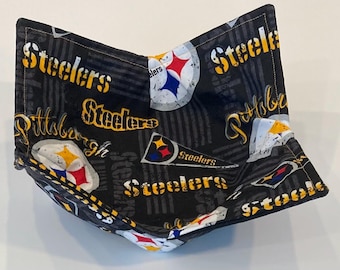 May include: A black and gray fabric bowl cozy with the words "Steelers" and "Pittsburgh" in yellow and white. The cozy features the Steelers logo in red, white, and blue. The bowl cozy is designed to keep food warm.