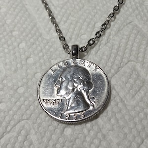 Może przedstawiać: Srebrny naszyjnik z zawieszką w postaci amerykańskiej ćwierćdolara z 1963 roku. Na monecie widnieje profil George'a Washingtona oraz napisy "Liberty", "In God We Trust" i "1963".